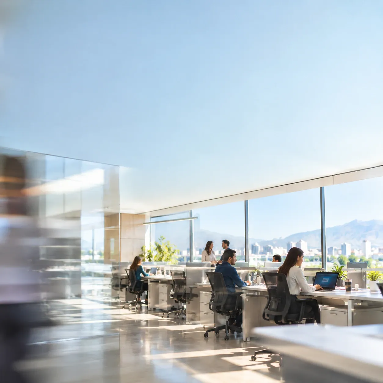 Open-plan office with teams working by floor-to-ceiling windows, city in the background
