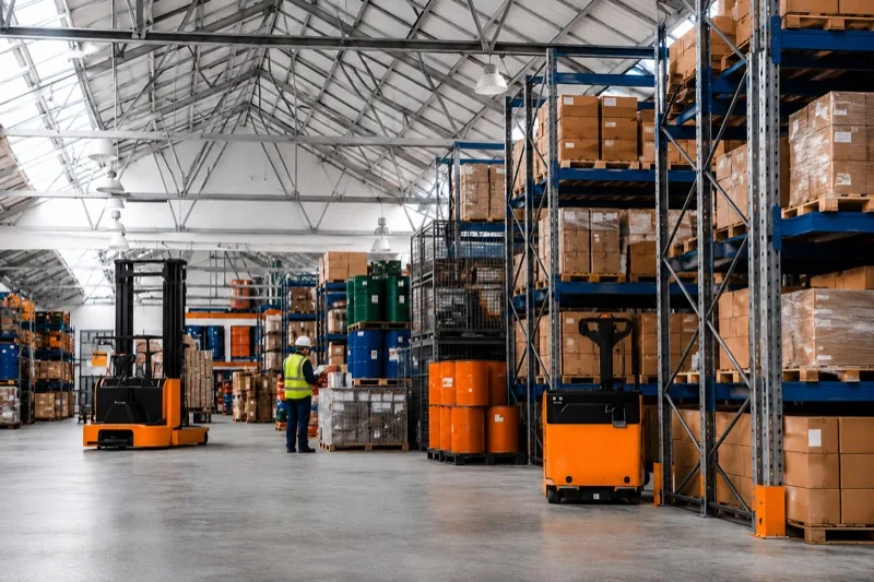 Almacén de distribución con racks de tarimas, montacargas eléctricos y un operador revisando inventario